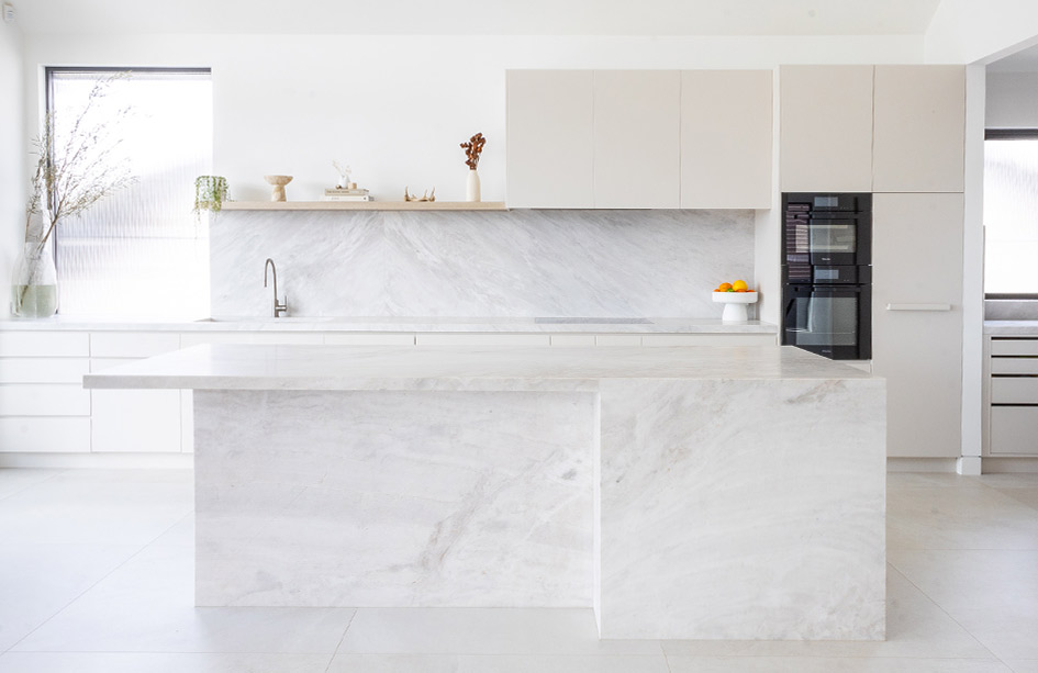 A modern kitchen featuring a Biancatto quartzite island with a stepped waterfall edge. The stone's soft grey veining pairs perfectly with warm matte cabinetry and a floating timber shelf, creating a calm, inviting space.