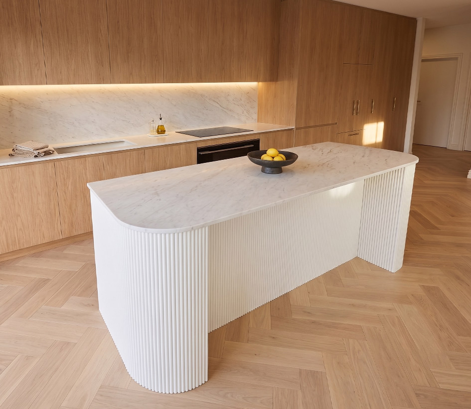 warm, modern kitchen featuring a curved Bianco Carrara marble island benchtop. The fluted white base has a distinct, non-monolithic shape, perfectly paired with warm timber cabinetry and herringbone floors for balance