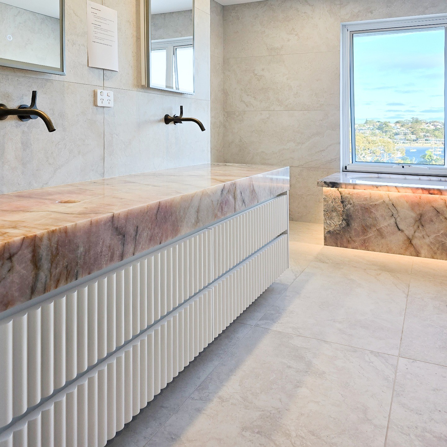 HERO. Pink quartzite bath surround, polished finish, mitred deck over an undermount bath with a backlit apron, Sydney harbour-view bathroom.