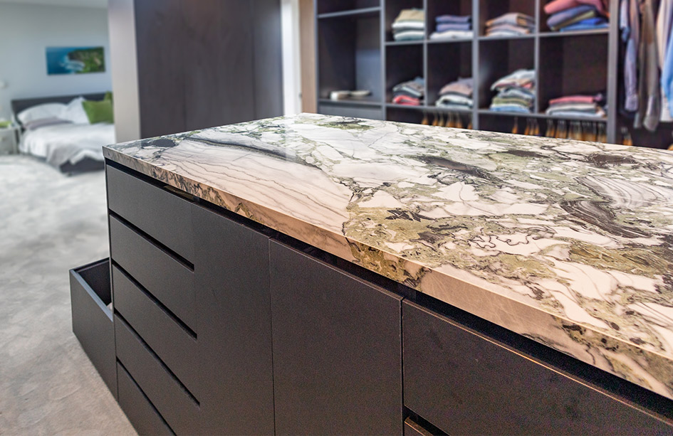 A luxurious walk-in closet featuring a custom dark island dresser topped with a striking Da Vinci Porcelain benchtop. The stone's dramatic green and black veining creates a bold centerpiece in this modern bedroom suite