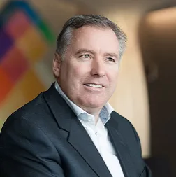 Helmuth Beck junior sitzt in einem Anzug vor einer farbenfrohen Wand.