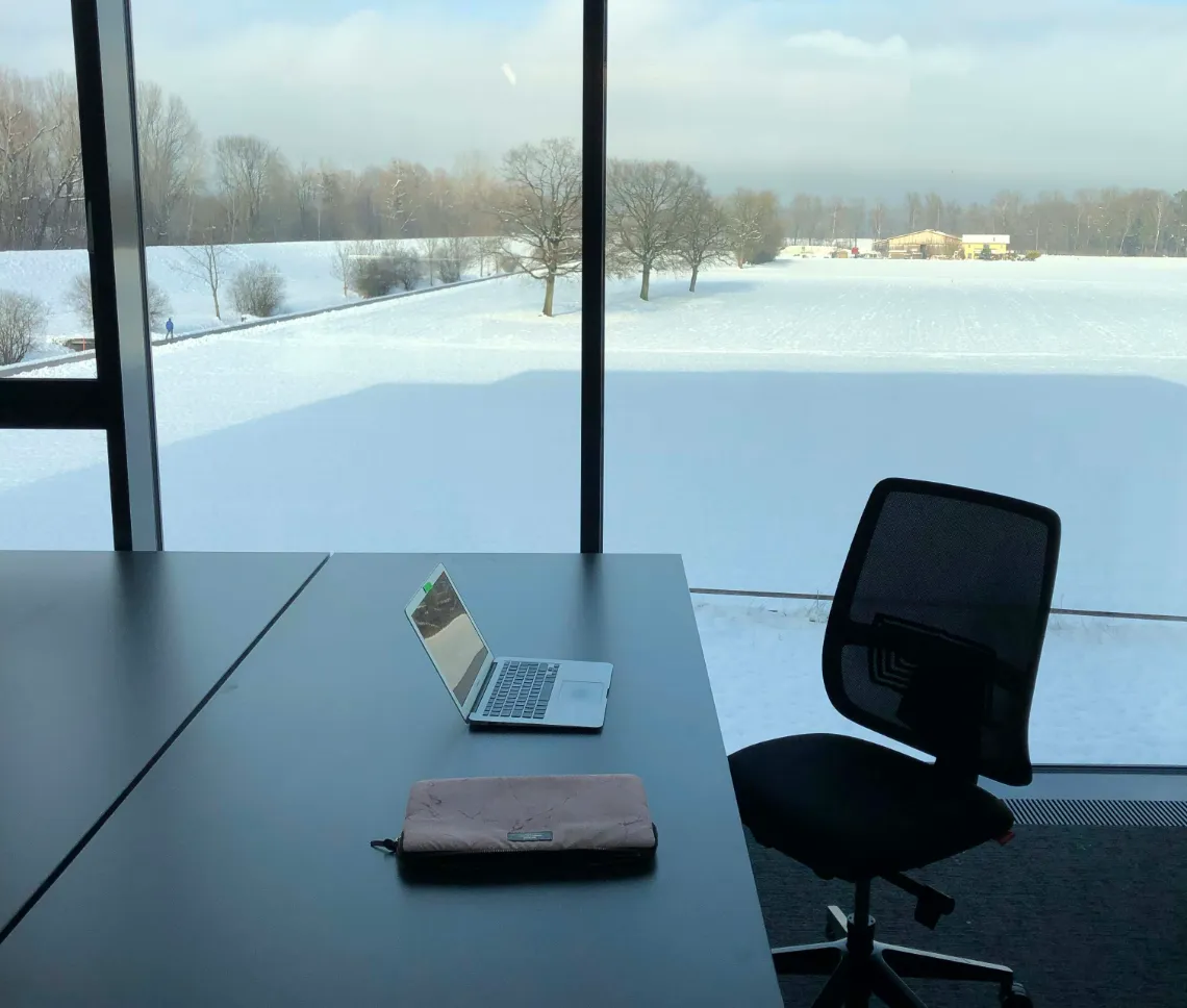Ein Laptop steht auf einem Schreibtisch vor einem Fenster, Schneelandschaft im Hintergrund.