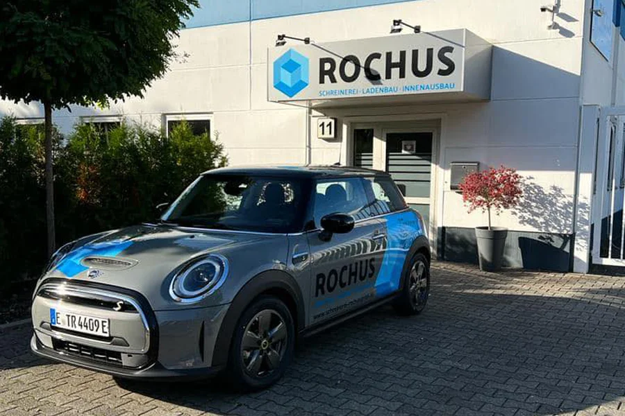Ein Firmenfahrzeug mit der Aufschrift „ROCHUS“ steht vor dem Eingangsbereich eines modernen Gebäudes mit dem Firmenschild „ROCHUS – Schreinerei · Ladenbau · Innenausbau“. Die Szene ist bei Tageslicht, mit gepflegter Bepflanzung und sauberem Pflasterweg.