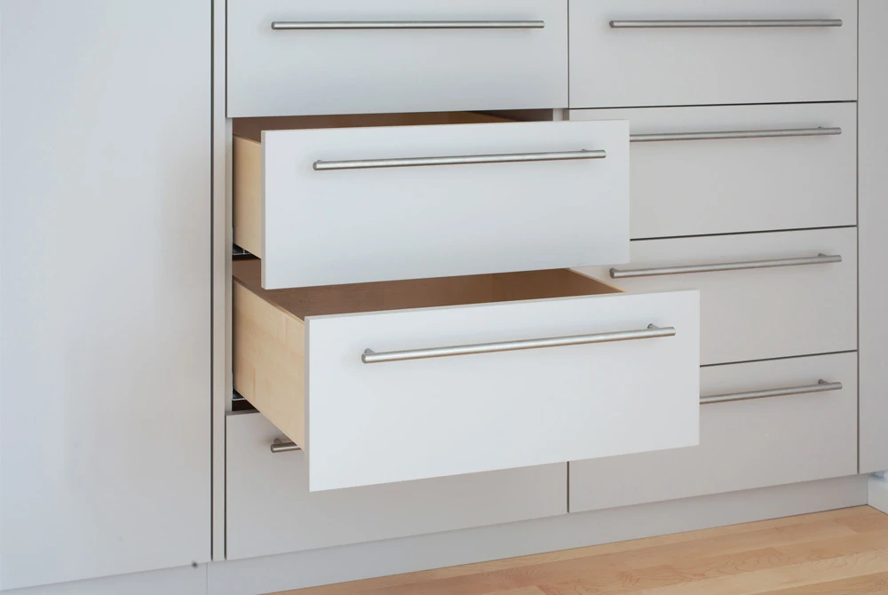 Weiße Schubladen mit langen, metallischen Griffleisten in einem modernen Schranksystem, teilweise geöffnet und auf hellem Holzboden stehend.