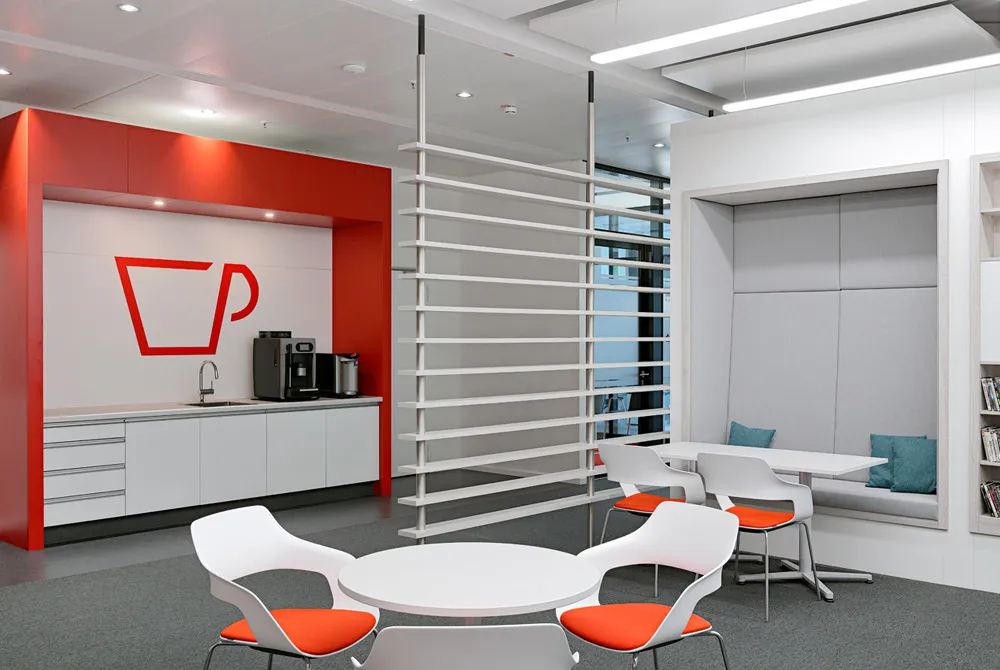 Moderne Teeküche in einem Büro mit weiß-rotem Design, Barhockern und integrierter Kaffeenische in einem hellen Raum mit Holzfußboden und Garderobe.