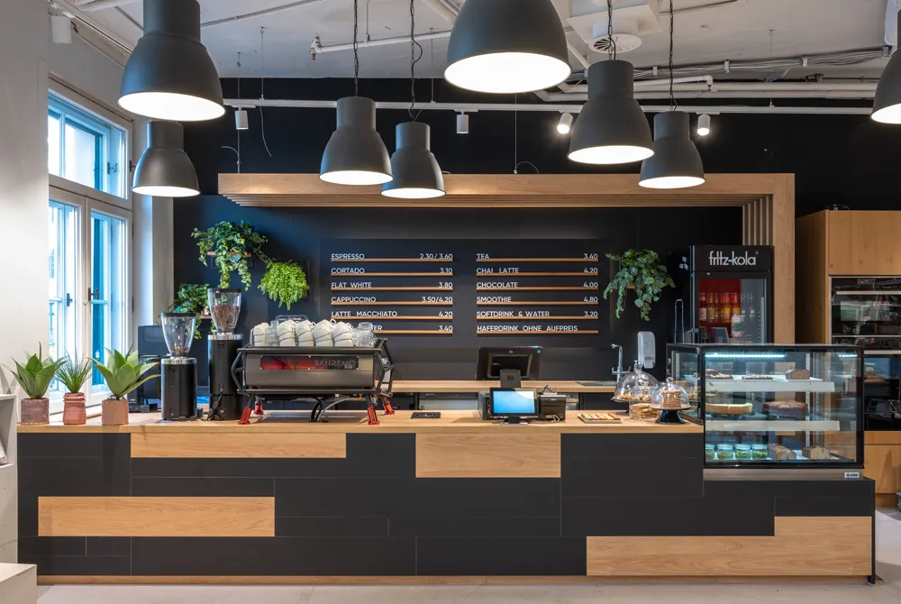 Stylischer Café-Tresen mit Holzverkleidung, großen Hängelampen und moderner Espressomaschine vor schwarzer Wand mit Menütafeln.
