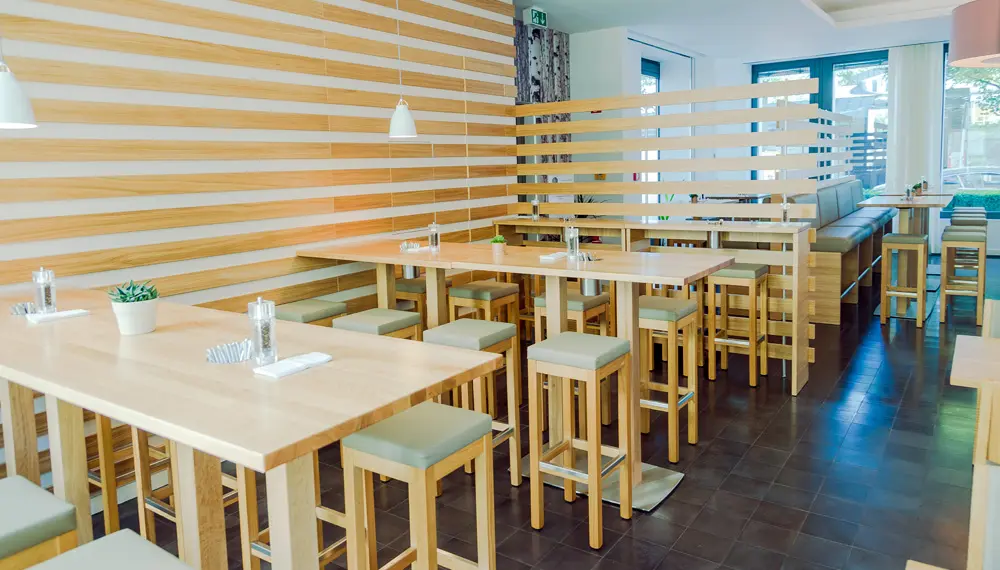 Moderne Kantine mit hellem Holzinterieur, hohen Tischen und Hockern, durch Holzlamellen strukturierter Raum, große Fenster für viel Tageslicht, freundliche Atmosphäre.
