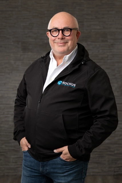Thomas Rochus steht lächelnd vor einem dunklen Hintergrund. Er trägt eine schwarze Jacke mit dem Logo „ROCHUS“ und eine weiße Hemdbluse.