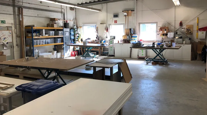 Helle und gut organisierte Werkstatt mit höhenverstellbaren Arbeitstischen, Werkzeugwänden und Lagerregalen.