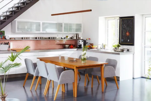 Helle Wohnküche mit Esstisch aus Holz, grauen Polsterstühlen, moderner Einbauküche und Kupferrückwand unter der Treppe.