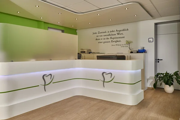 Heller Empfangsbereich einer Zahnarztpraxis mit modern geschwungener weißer Theke, grünen Akzenten und Zahn-Logo. Im Hintergrund ein Goethe-Zitat an der Wand, daneben Pflanzen und Bürotresen mit Geräten. Freundliche, einladende Atmosphäre.