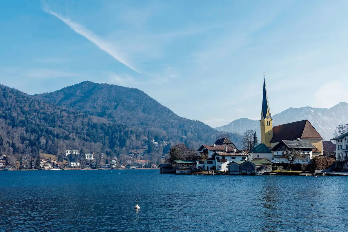 Idyllische Seelandschaft in Bayern: Im Vordergrund der Tegernsee mit einem Schwan, im Hintergrund ein malerisches Dorf mit Kirche und spitzem Kirchturm vor bewaldeten Alpenhängen.