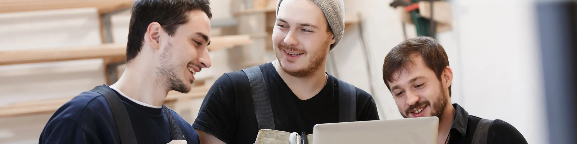 Drei junge Handwerker in Arbeitskleidung lachen gemeinsam über etwas auf einem Laptop in einer Werkstattumgebung.