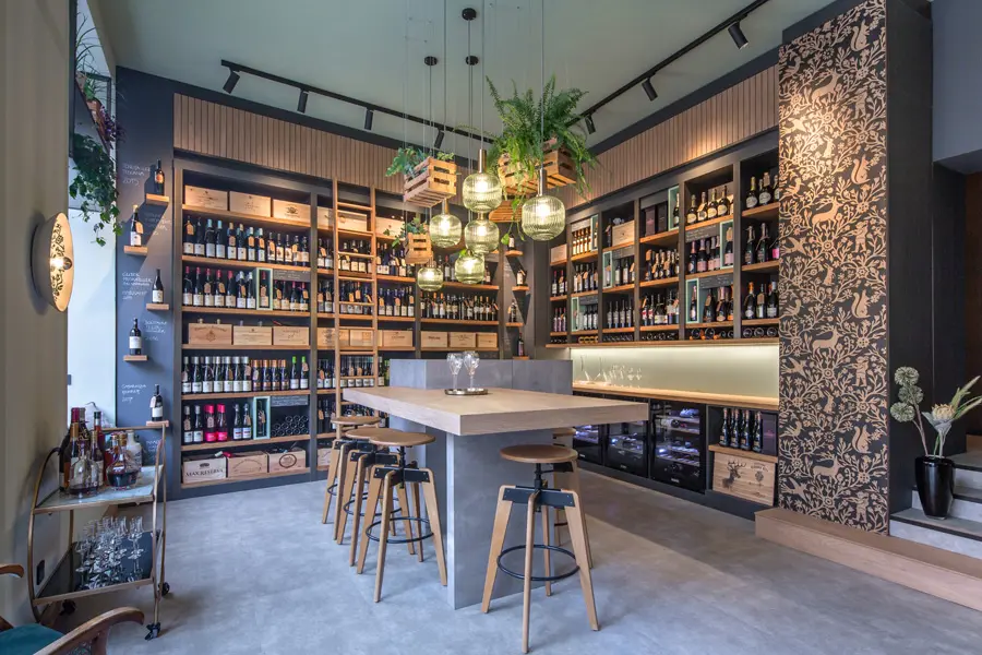 Modernes Weinatelier mit hohen Weinregalen, zentralem Verkostungstisch, Barhockern und hängenden Pflanzen unter stilvollen Lampen.