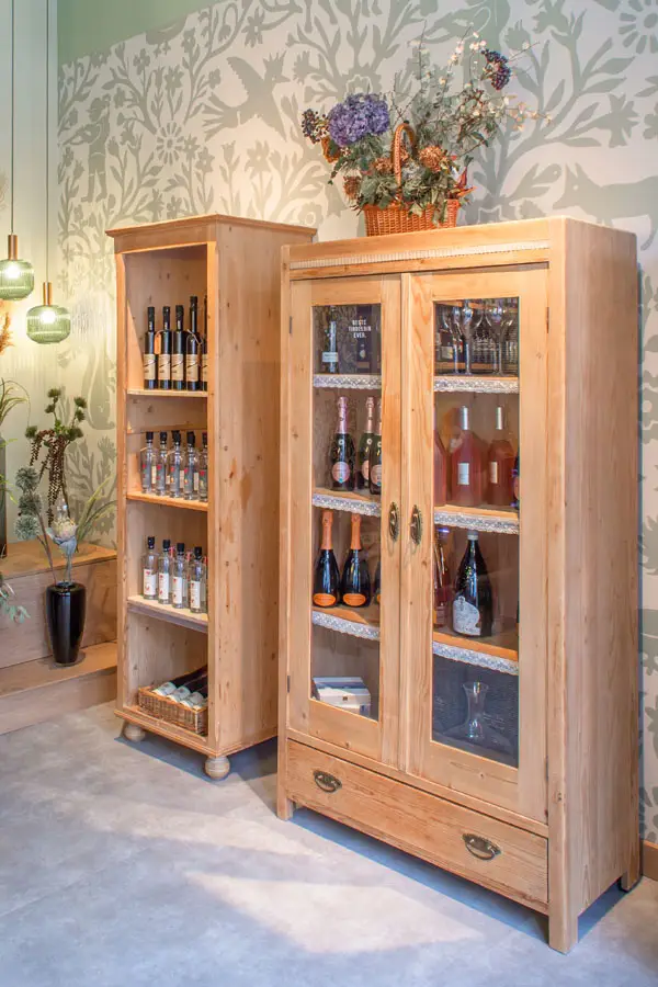 Rustikale Holzvitrinen im Weinatelier Rochus mit erlesenen Spirituosen und Sektflaschen vor floraler Wandgestaltung.