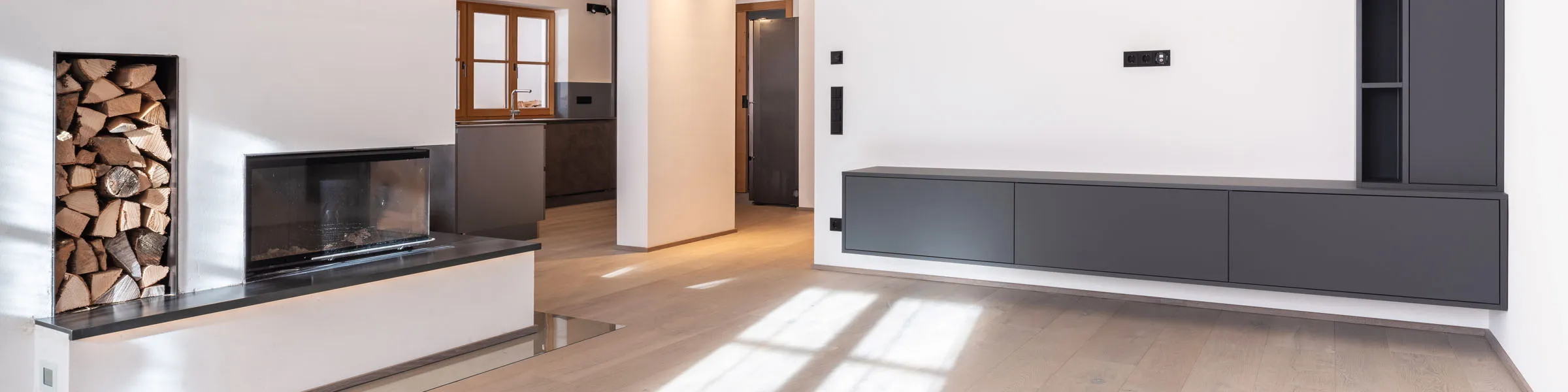 Modernes Wohnzimmer mit Kamin, Holzlager, dunklem Wandboard und Blick in eine offene Küche mit Designermöbeln.