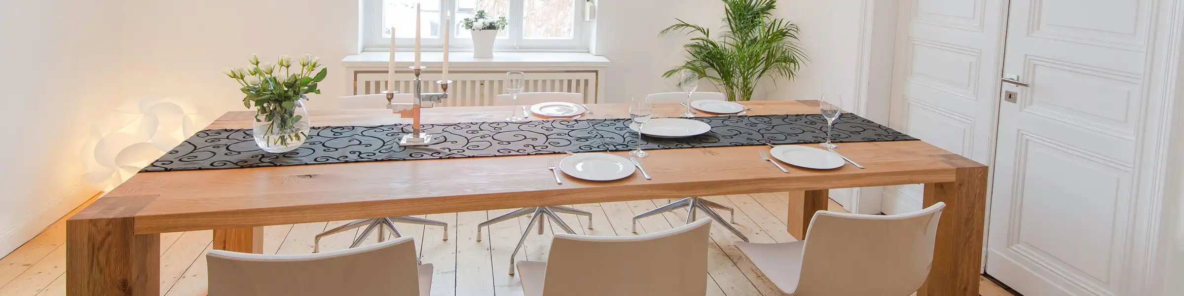 Moderner Essbereich mit großem Holztisch, sechs weißen Drehstühlen und stilvoller Tischdekoration in einem hellen Raum mit Holzdielen und hohen Fenstern.