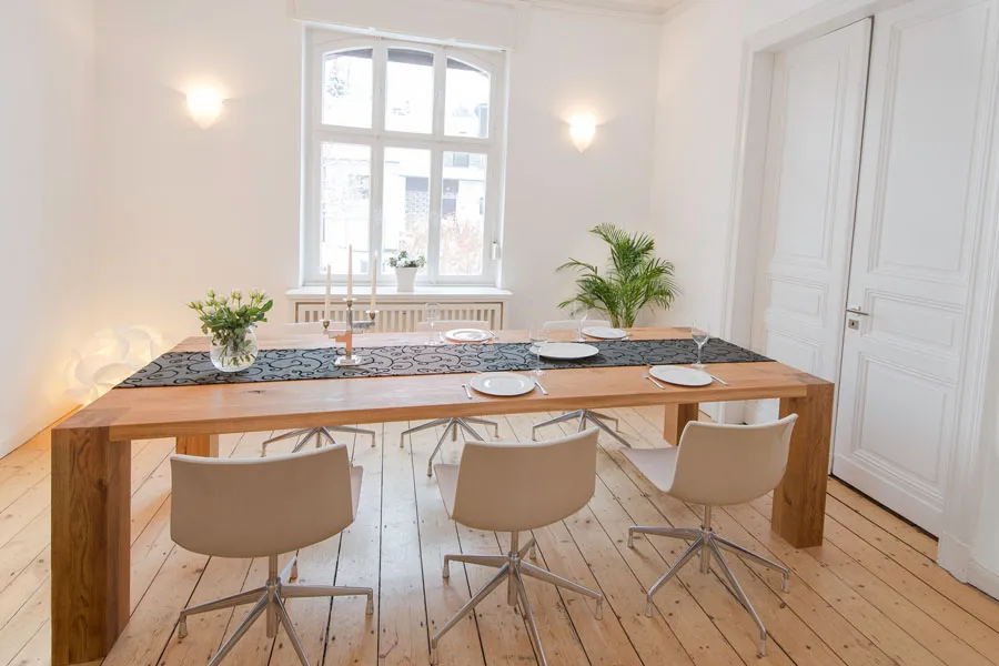 Moderner Essbereich mit großem Holztisch, sechs weißen Drehstühlen und stilvoller Tischdekoration in einem hellen Raum mit Holzdielen und hohen Fenstern.