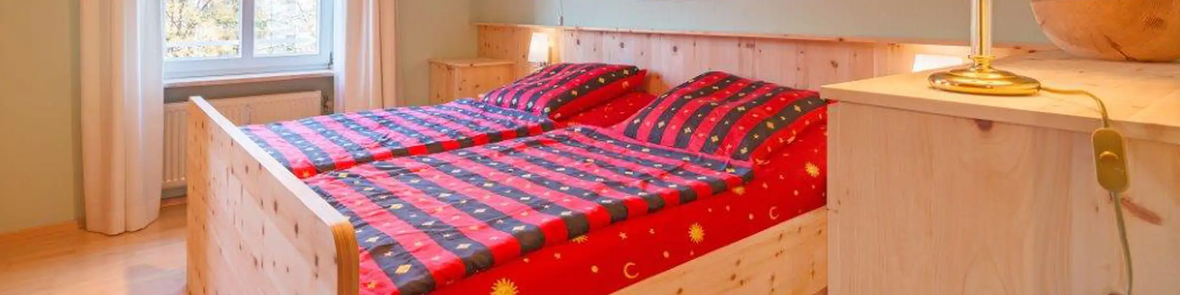 Gemütliches Schlafzimmer mit hellem Zirbenholzbett, roter Bettwäsche mit Sonnenmotiv und warmer Beleuchtung in traditionellem Stil.