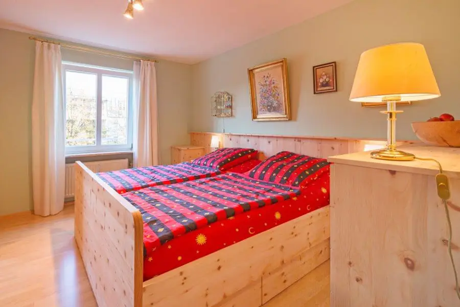 Gemütliches Schlafzimmer mit hellem Zirbenholzbett, roter Bettwäsche mit Sonnenmotiv und warmer Beleuchtung in traditionellem Stil.