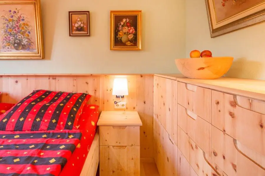 Detailansicht eines gemütlichen Schlafzimmers mit Nachttisch und maßgefertigter Kommode aus Zirbenholz, roter Bettwäsche und stimmungsvoller Wandbeleuchtung.