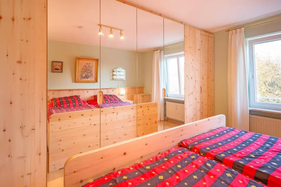 Schlafzimmer mit Zirbenholzbett, großem Spiegelschrank und heller Tageslichtatmosphäre durch zwei Fenster; rote Bettwäsche setzt farbliche Akzente.