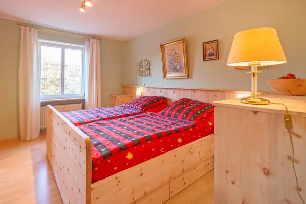 Gemütliches Schlafzimmer mit hellem Zirbenholzbett, roter Bettwäsche mit Sonnenmotiv und warmer Beleuchtung in traditionellem Stil.