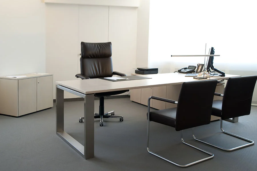 Modernes Chefzimmer mit elegantem Schreibtisch, ergonomischem Leder-Bürostuhl und zwei Besucherstühlen – minimalistisches Design bei natürlichem Tageslicht.