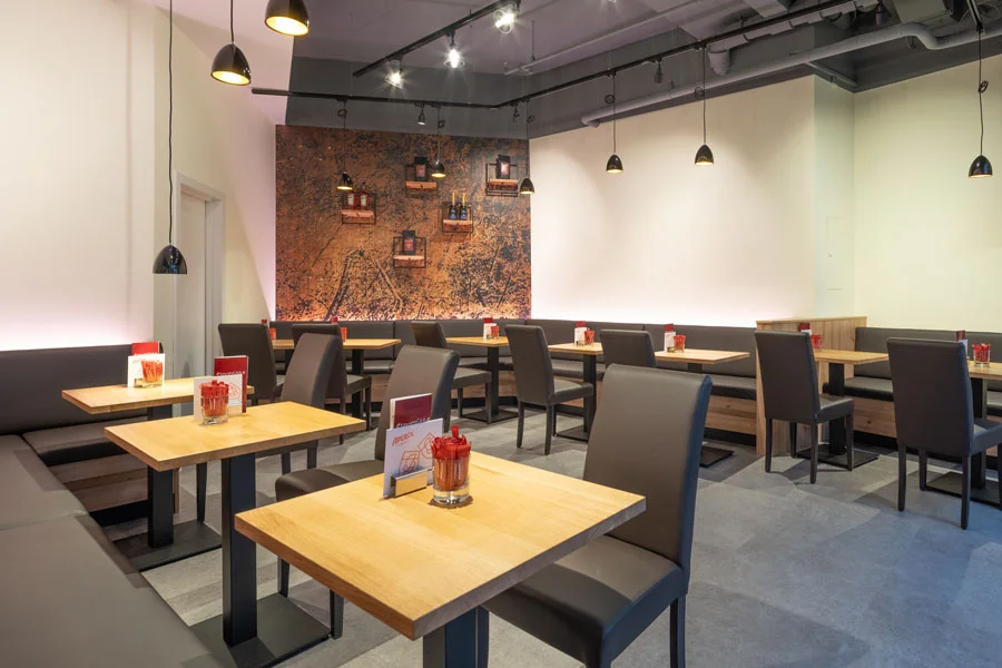 Modern eingerichtetes Restaurant mit hellen Holztischen, dunklen Polsterbänken und Designerleuchten – gemütliches Ambiente mit stilvoller Wandgestaltung.