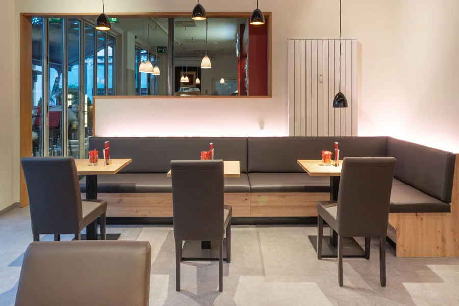 Gemütliche Restaurant-Sitzecke mit gepolsterter Eckbank, Holztischen und modernen Pendelleuchten – einladende Atmosphäre mit Blick in den Gastraum durch ein großes Innenfenster.