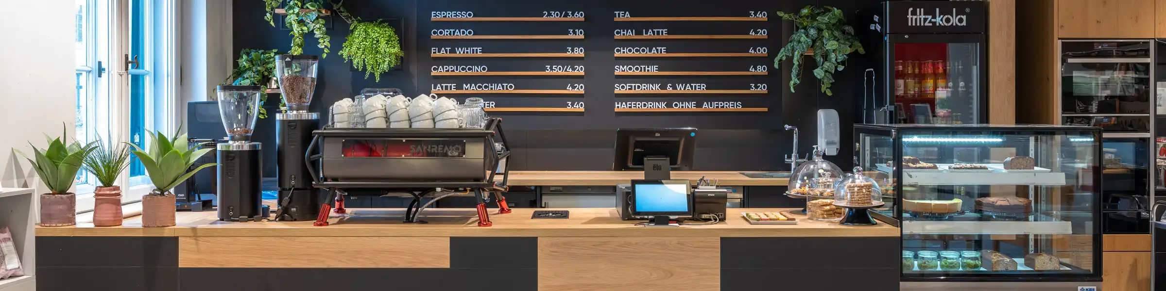 Stylischer Café-Tresen mit Holzverkleidung, großen Hängelampen und moderner Espressomaschine vor schwarzer Wand mit Menütafeln.