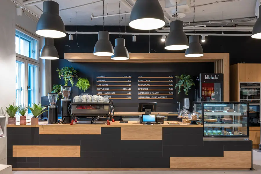 Stylischer Café-Tresen mit Holzverkleidung, großen Hängelampen und moderner Espressomaschine vor schwarzer Wand mit Menütafeln.