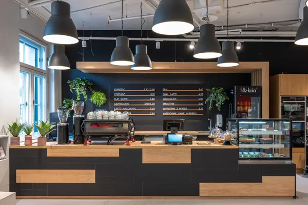 Stylischer Café-Tresen mit Holzverkleidung, großen Hängelampen und moderner Espressomaschine vor schwarzer Wand mit Menütafeln.