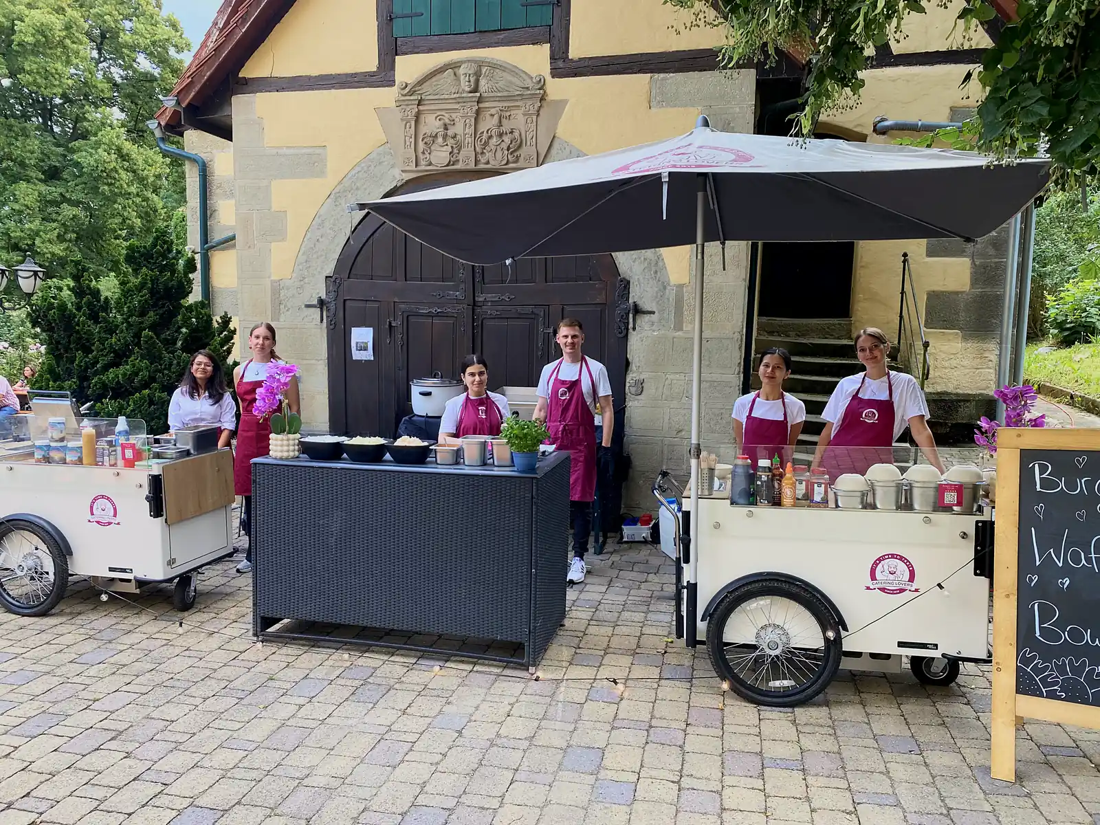 Zwei CateringLovers-Stationen mit Crew und Kreidetafel im Innenhof vor einem historischen Torbogen