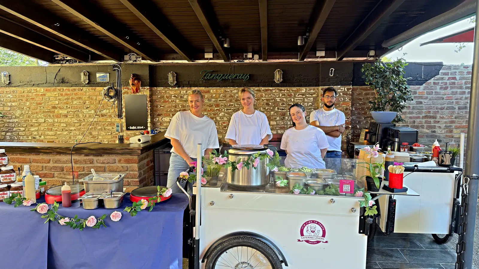 CateringLovers-Station und Ausgabetisch in einer überdachten Backsteinhalle, vier Crewmitglieder dahinter