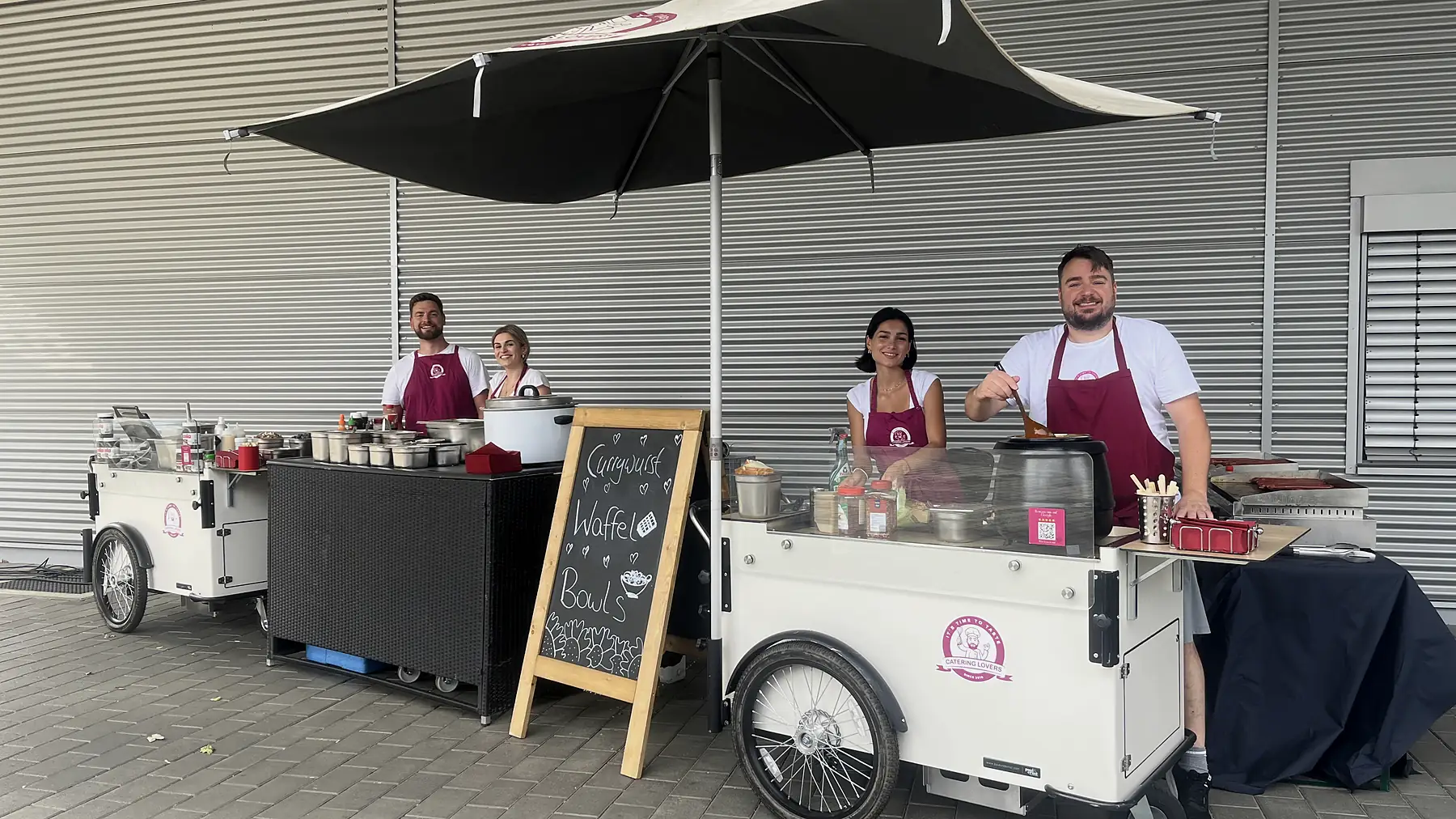 Funktionierende Station: vier CateringLovers-Mitarbeiter an zwei HappyBike-Wagen mit Kreidetafel Currywurst, Waffeln und Bowls.