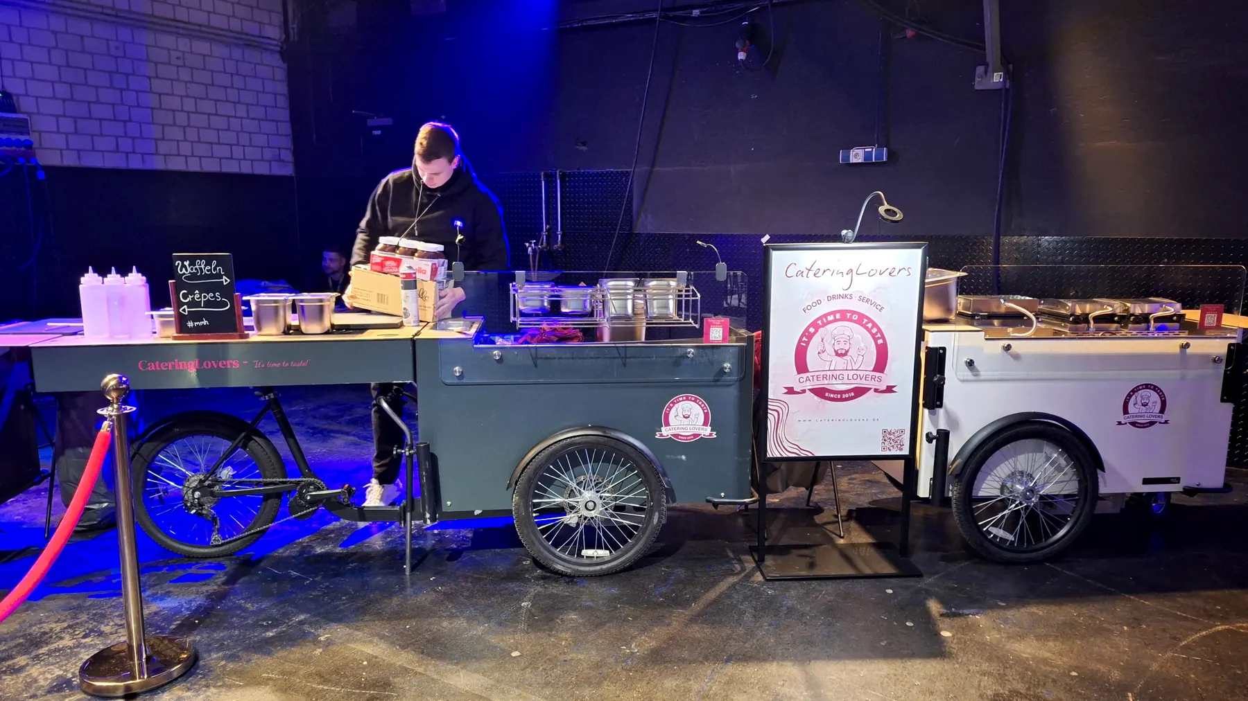Aufbau der Catering-Station in der noch dunklen Messehalle zwischen Absperrkordel und Kartons