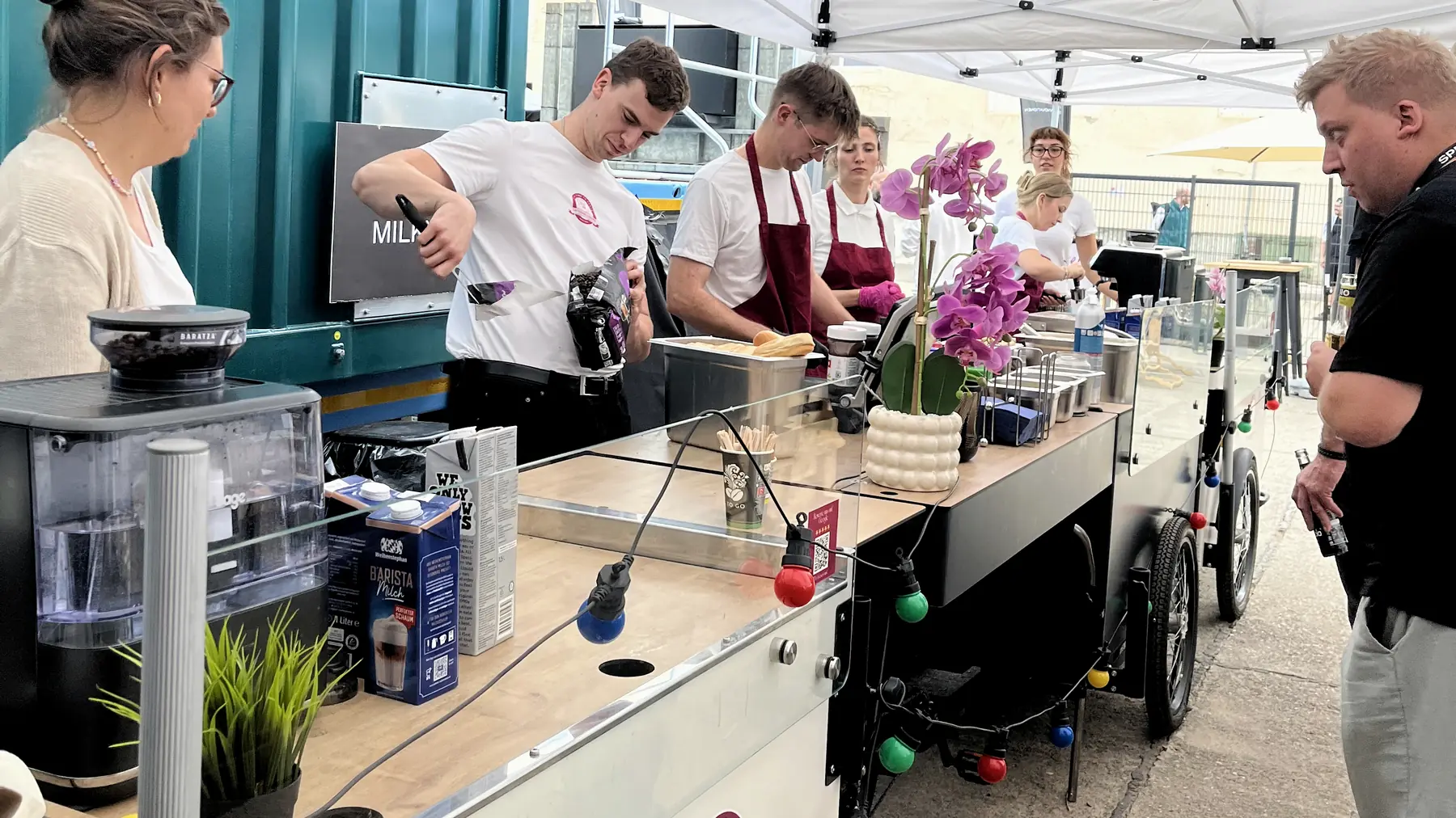 Laufende Kaffeestation auf dem Messegelände: Barista am Siebträger und Mühle, Kollegen bereiten Speisen zu, ein Gast wartet am Tresen