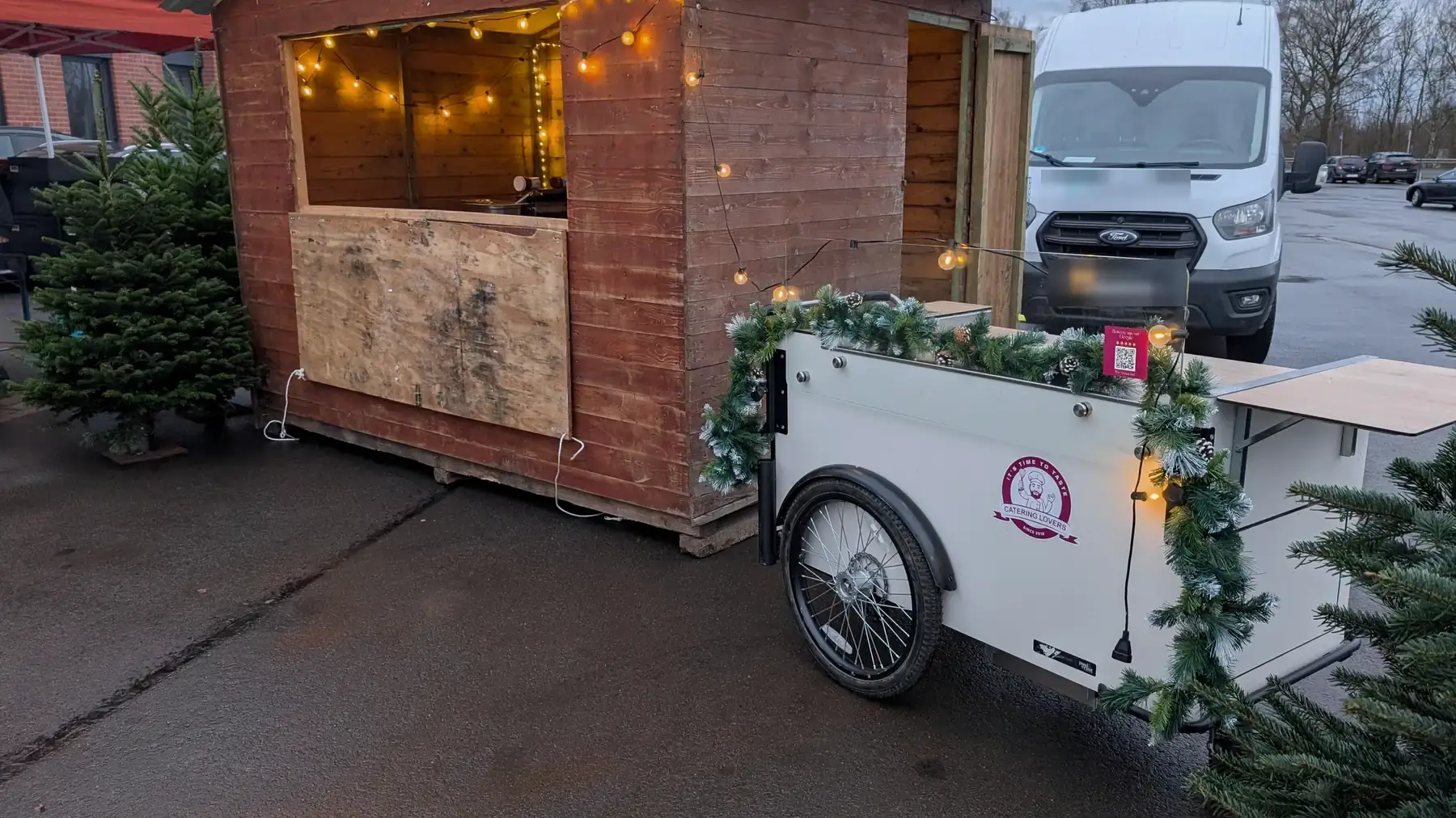 Firmen-Weihnachtsmarkt im Aufbau: Holzhütte mit Lichterkette, davor ein weißes HappyBike mit Tannengirlande, links und rechts echte Nordmanntannen