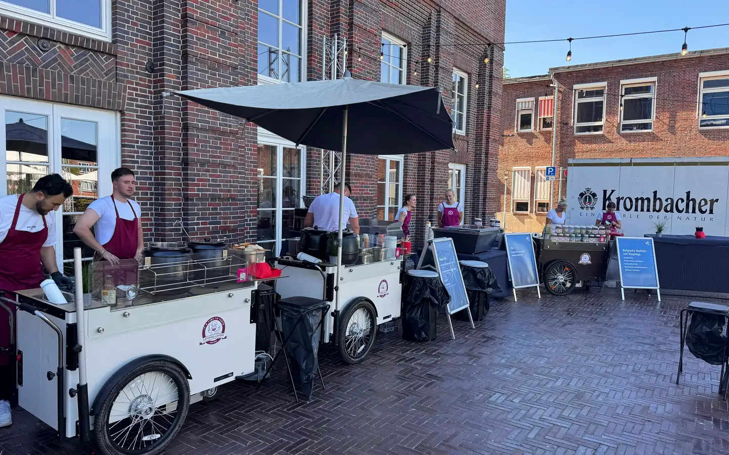 Mehrere HappyBike-Stationen in einer Reihe auf einem Innenhof, Crew in weinroten Schürzen arbeitet an den Grillplatten