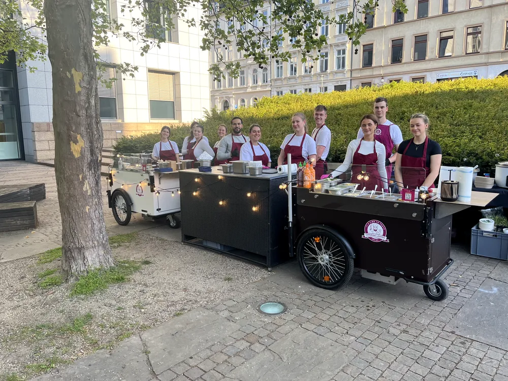 Elfköpfige Crew in weißen Shirts und weinroten Schürzen hinter zwei aufgebauten Stationen in einem Stadtpark
