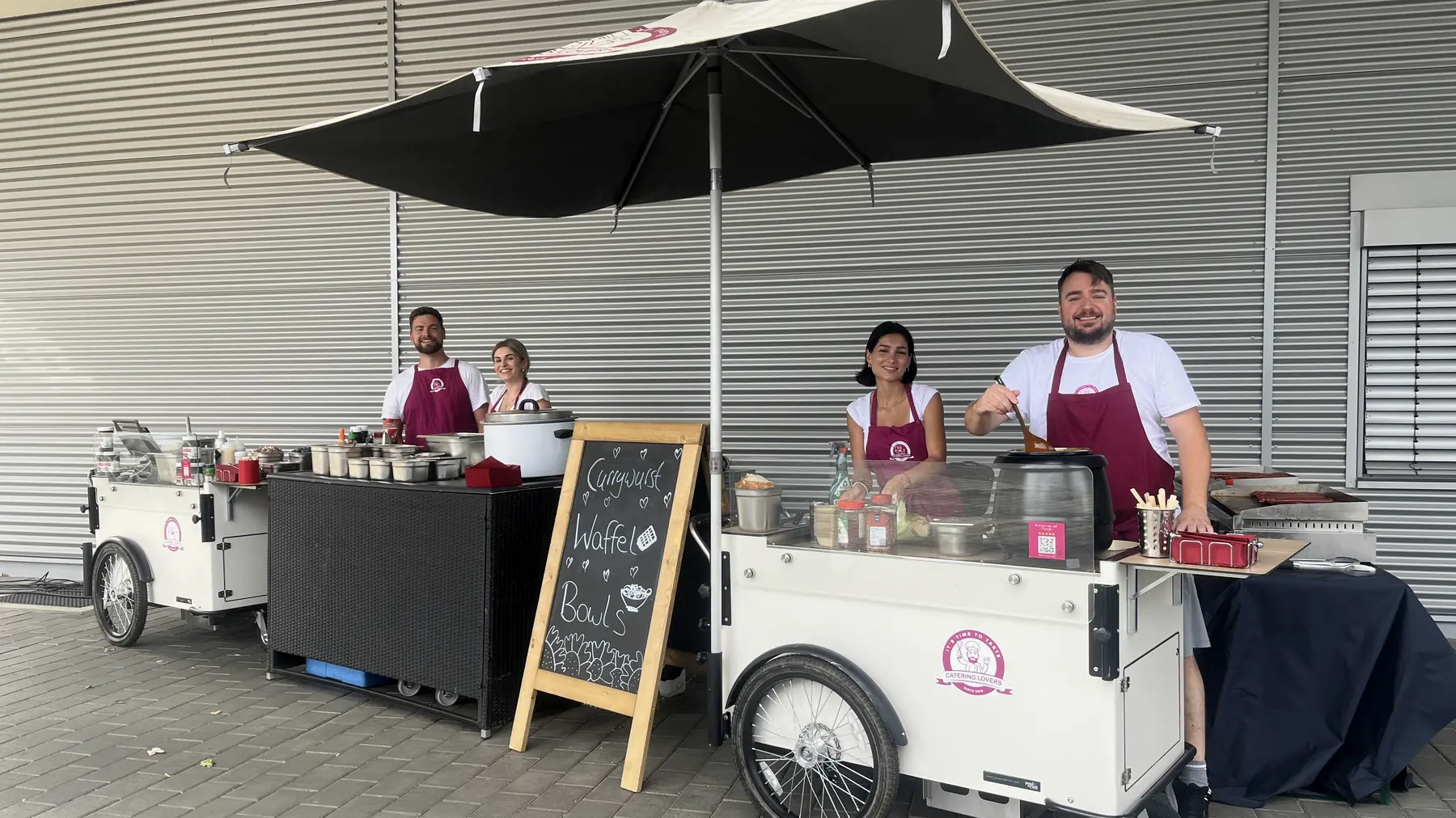 Zwei identisch aufgebaute HappyBike-Stationen mit Sonnenschirm, Kreidetafel und vier Crew-Mitgliedern vor einer Industriehalle