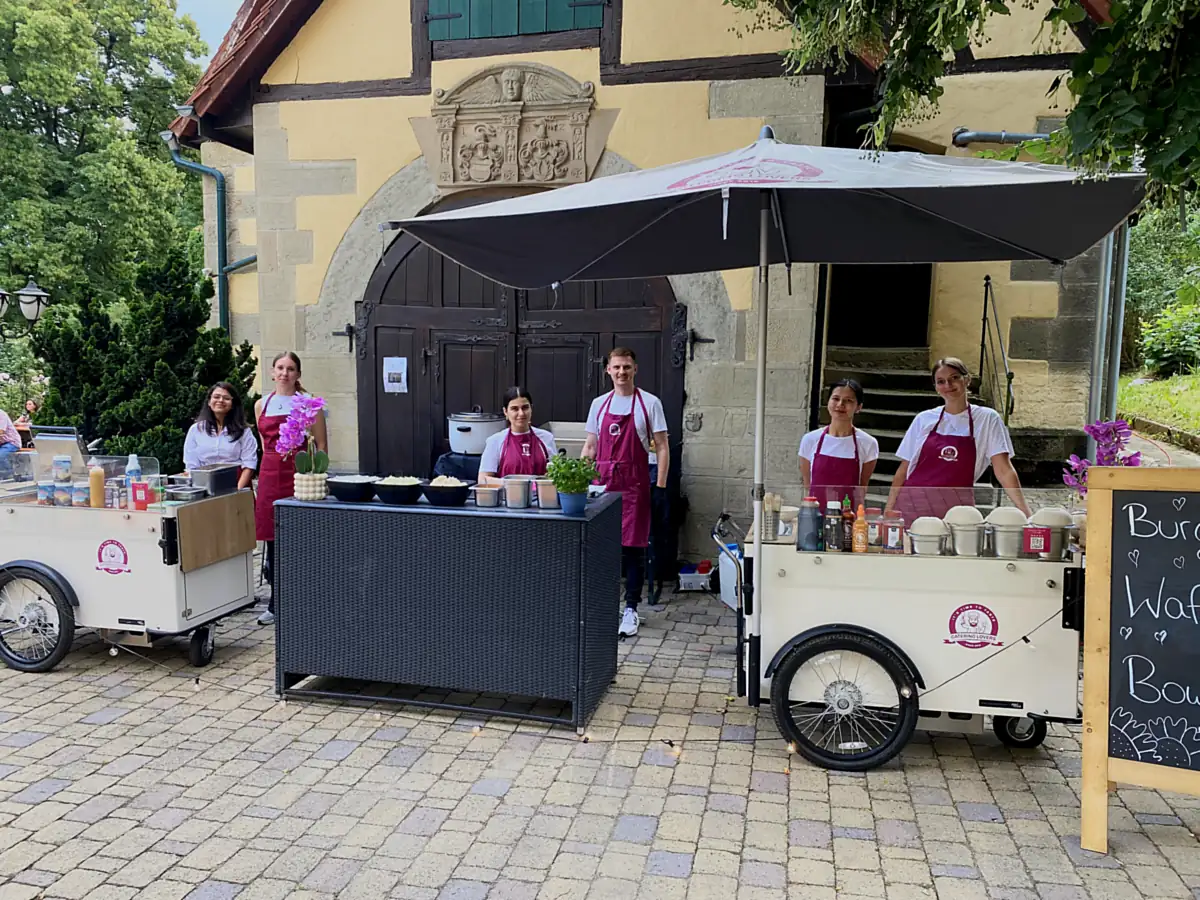 Sechs Crew-Mitglieder mit zwei HappyBikes und Ausgabetresen vor einem historischen Torhaus auf gepflastertem Platz