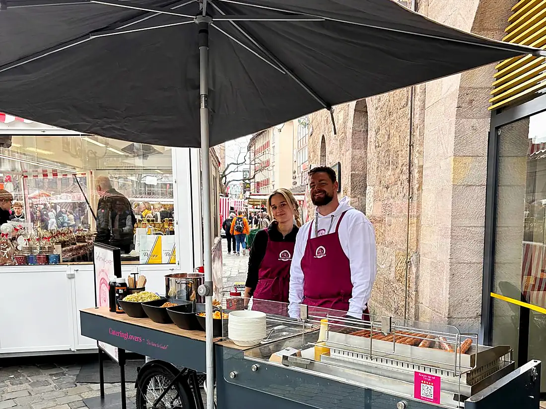 HappyBike-Station mit zwei Crew-Mitgliedern in einer Fußgängerzone neben einem Marktstand, im Hintergrund Passanten und ein historischer Torbogen