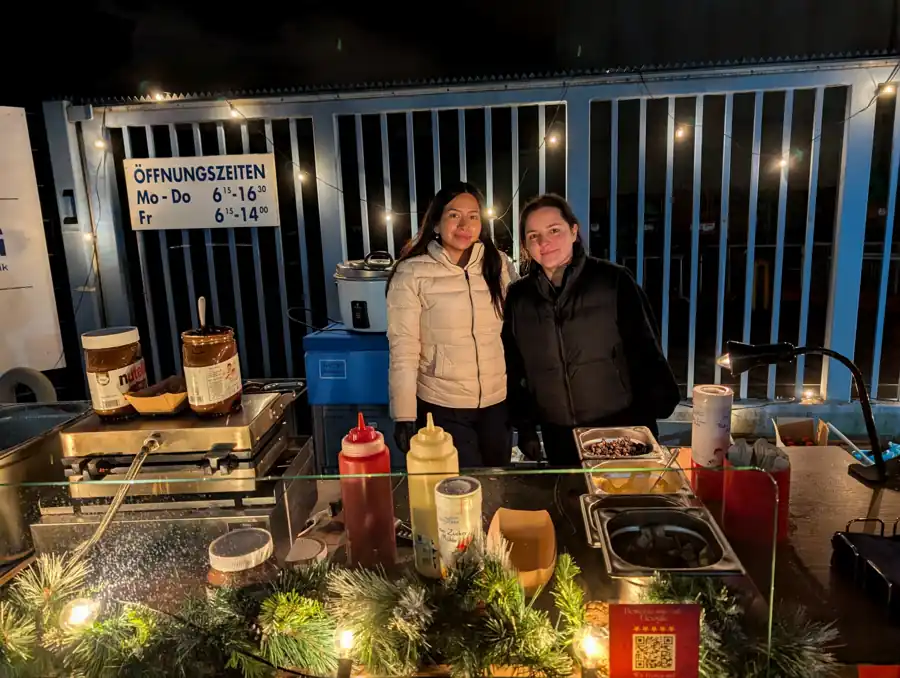Nachts an der beleuchteten Waffelstation: zwei Gäste in Winterjacken an der Ausgabe, dahinter Lichterkette und Werkstor