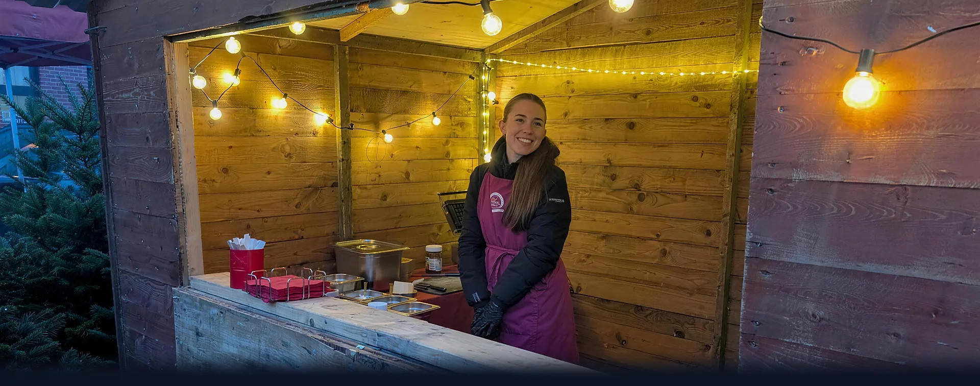Weihnachtsmarkt-Holzhütte von CateringLovers mit warmer Lichterkette: Gastgeberin lächelt hinter dem Tresen in der Abenddämmerung.