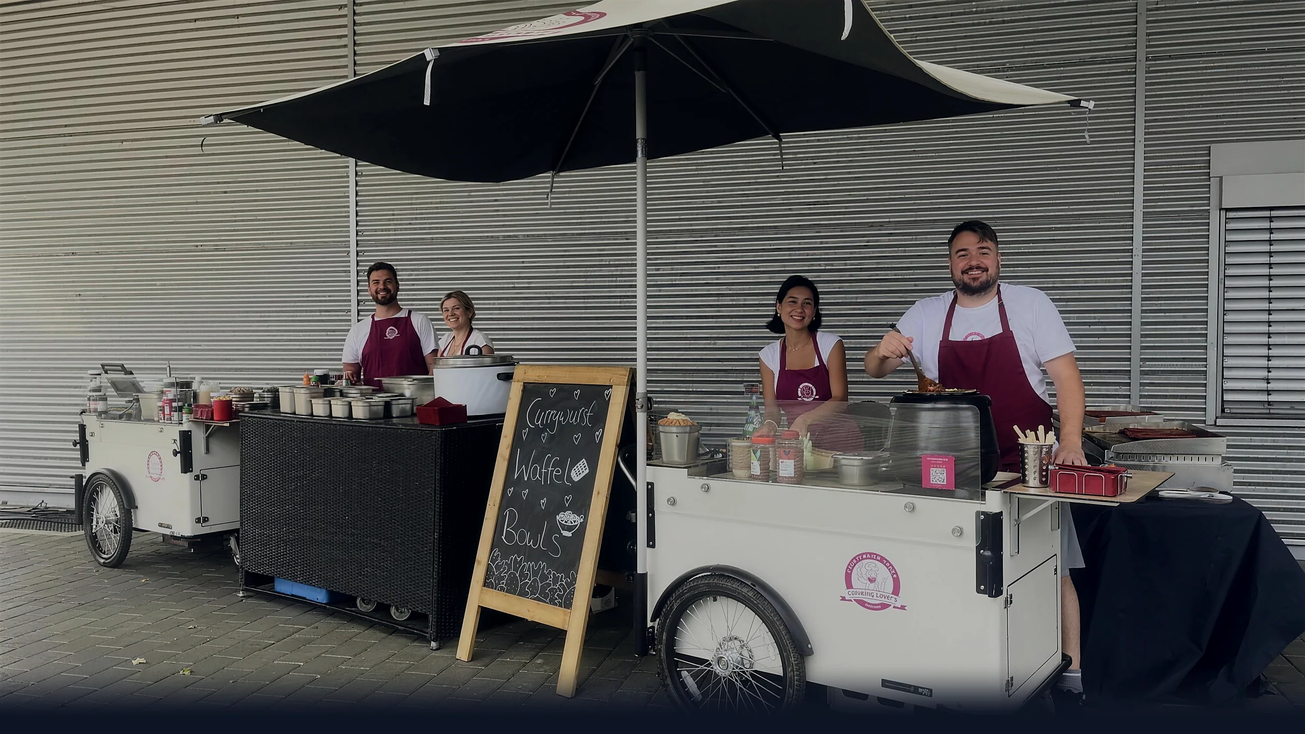 Zwei aufgebaute Live-Cooking-Stationen von CateringLovers mit vier Crew-Mitgliedern und Kreidetafel vor einer Firmenfassade