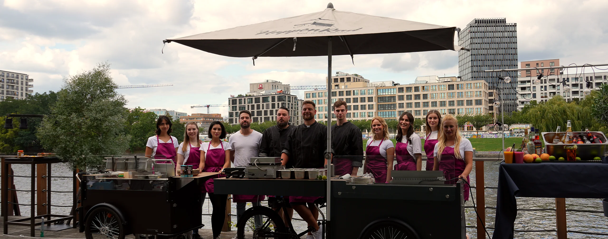CateringLovers-Crew am HappyBike-Verbund unter dem Schirm, dahinter Fluss und Skyline