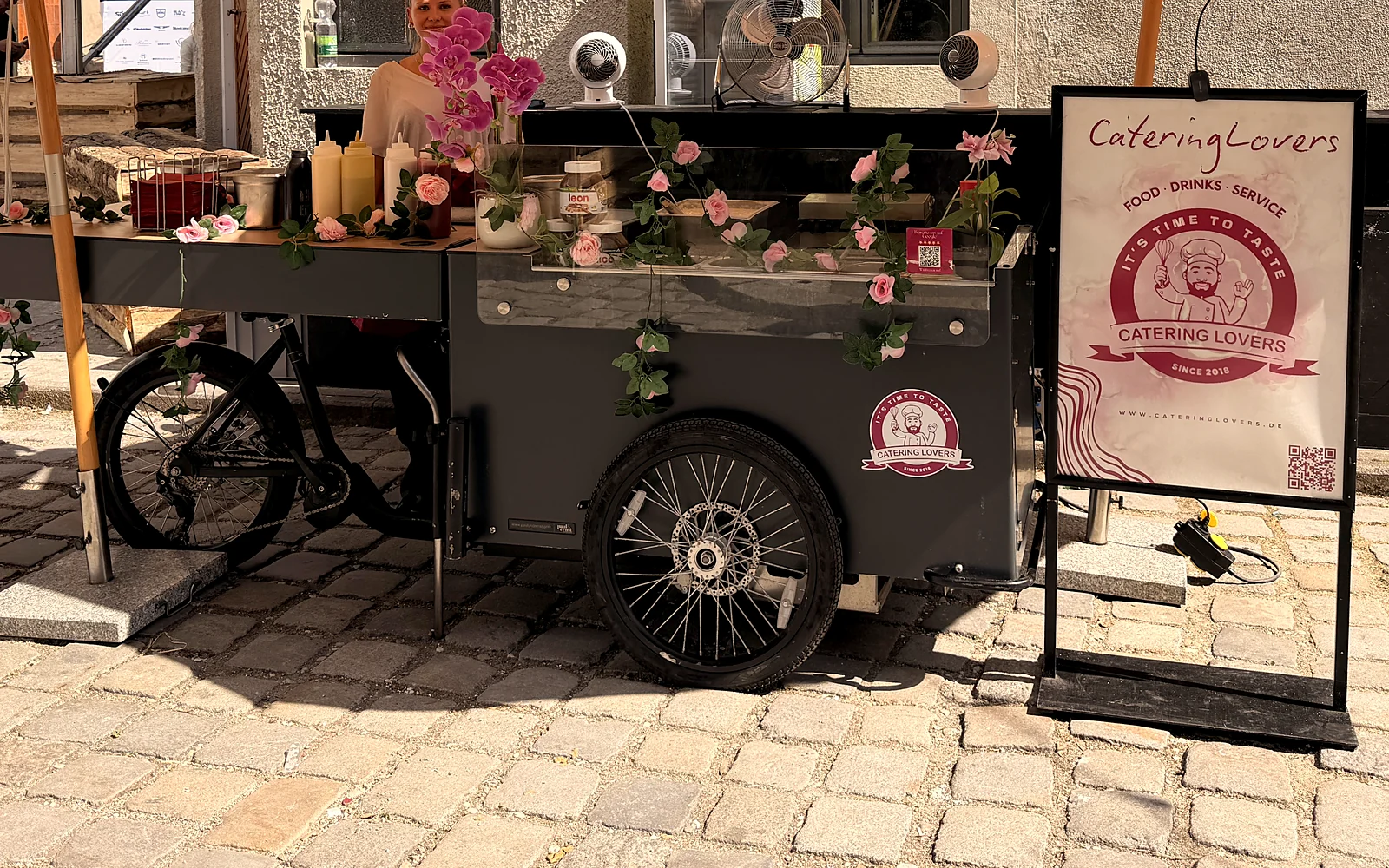 HappyBike Big: dunkelgraues Lastenrad mit ausgeklappter Theke und Rosen-Deko unter Sonnenschirmen auf einem Altstadthof