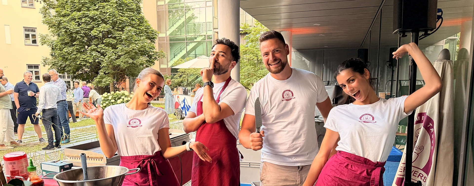 Sommerfest im Firmenhof: vier Crew-Mitglieder scherzen an der Station, dahinter Gäste im Grünen
